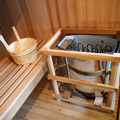the wooden Cask & ladle on the bottom bench and the silver Harvia 6.0 kW heater in a Wood inside of the protective wood safety guard next to the bottom bench in the Charleston 4-person traditional sauna in hemlock, double bench,