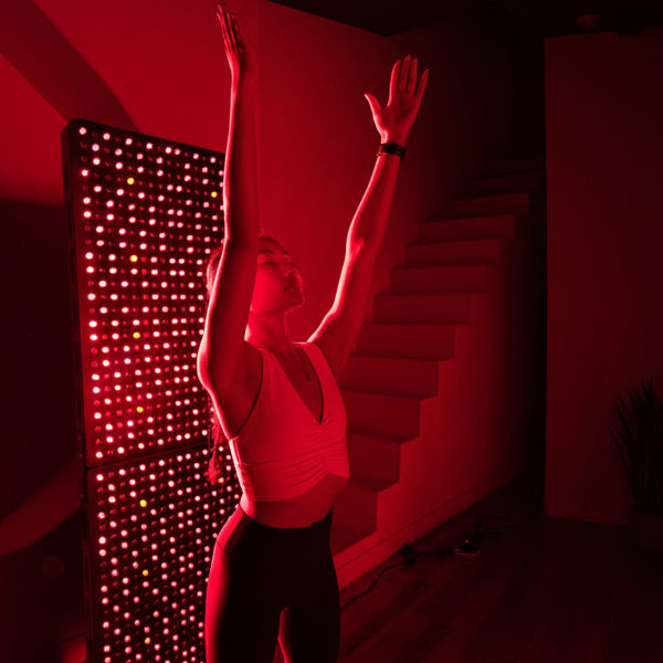 Woman in a white tank top and black leggings standing with arms raised in front of a red light therapy panel.